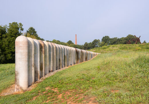 A long, straight line of round-topped cylindrical vessels that appear to recede into a hillside. In the background there are trees, a smoke stack, and a clear blue sky.