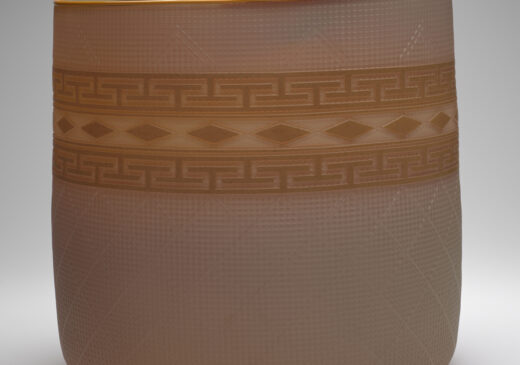 A glass basket set against a gray-toned backdrop. The basket is a semi-transparent cream color, with a golden-yellow band of design around the middle. The rim of the basket is also golden yellow. The design band is made up of a top and bottom band of repeating geometric shapes. There is a row of repeating diamond shapes between the geometric shapes.