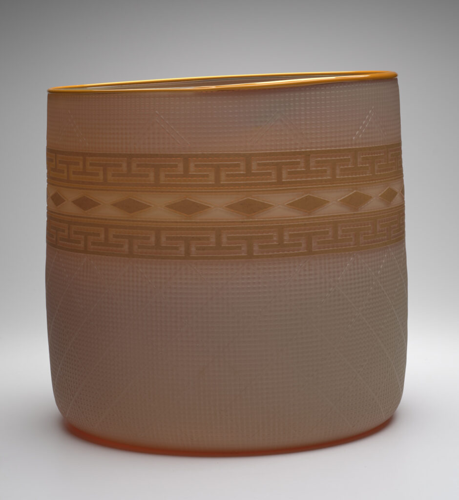 A glass basket set against a gray-toned backdrop. The basket is a semi-transparent cream color, with a golden-yellow band of design around the middle. The rim of the basket is also golden yellow. The design band is made up of a top and bottom band of repeating geometric shapes. There is a row of repeating diamond shapes between the geometric shapes.