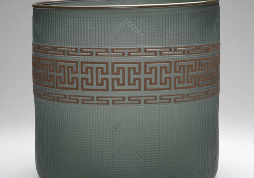 A glass basket set against a gray-toned backdrop. The basket is a semi-transparent dark green color, with a brown design band wrapped around the middle. The design band is made up of an identical top and bottom row, with an angular repeating pattern design. The middle row features repeating geometric shapes.