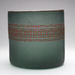 A glass basket set against a gray-toned backdrop. The basket is a semi-transparent dark green color, with a brown design band wrapped around the middle. The design band is made up of an identical top and bottom row, with an angular repeating pattern design. The middle row features repeating geometric shapes.