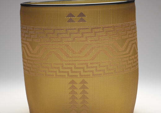 A glass basket set against a gray-toned backdrop. The basket is made of semi-transparent yellow glass, with brown designs etched into its surface. The horizontal designs wrap around the middle of the basket. The middle band is a geometric wave shape that is made up of three rows of lines. On either side of the middle band are identical bands of a repeating diagonal zig-zag shape. The main design is made up of two vertical columns of triangles. There are four triangles above the horizontal design bands, and there are 18 triangles below them.