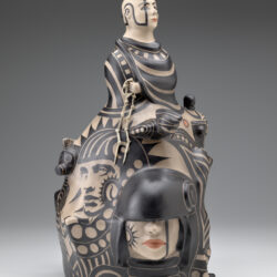 A ceramic sculpture against a gray-toned backdrop. The sculpture has a light beige base, with designs and figures painted in black and muted red-orange. The top of the sculpture is a bust of a man. His hair is up in buns with the sides of his head shaved bare, and he is wearing a cloak painted with stripes and squares. Flowing down from him on the base of the sculpture are 2D paintings and 3D protruding figures. These figures are a mix of men and women. They are dressed in different kinds of masks and helmets.