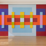 An installation made up of geometric squares and rectangles in a variety of sizes. Five of the rectangles are three-dimensional shapes that stick out slightly from the wall. They are lined up in a row through the middle of the mural. They are evenly spaced, with the middle rectangle slightly wider than the other four. The shapes are different shades of red, blue, orange, pink, yellow, and green.