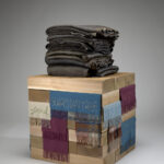 An image of a sculpture set against a gray-toned backdrop. The sculptural work features a stack of folded blankets made of bronze metal. The blankets are on top of a plinth made of wooden boards that are stacked in alternating directions. There are colorful pieces of wool blankets tucked in between the wooden boards. The blanket pieces are embroidered with words and phrases and have fringed edges.