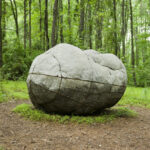 A boulderlike sculpture resembling a human figure curled up on the ground in a forest