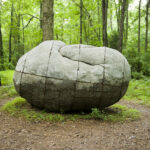 A boulderlike sculpture resembling a human figure curled up on the ground in a forest