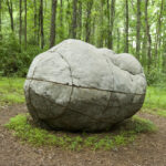 A boulderlike sculpture resembling a human figure curled up on the ground in a forest
