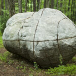 A boulderlike sculpture resembling a human figure curled up on the ground in a forest