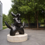 A dark brown sculpture of a birdlike creature on a concrete pedestal in an outdoor setting