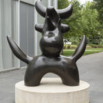 A dark brown sculpture of a birdlike creature on a concrete pedestal in an outdoor setting