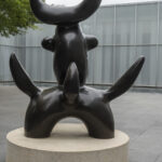 A dark brown sculpture of a birdlike creature on a concrete pedestal in an outdoor setting