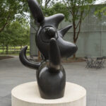 A dark brown sculpture of a birdlike creature on a concrete pedestal in an outdoor setting