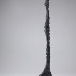 A bronze sculpture of an elongated nude female figure with oversized feet