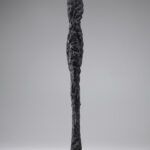 A bronze sculpture of an elongated nude female figure with oversized feet