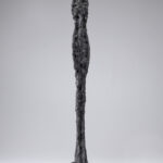 A bronze sculpture of an elongated nude female figure with oversized feet
