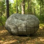 A boulderlike sculpture resembling a human figure curled up on the ground in a forest