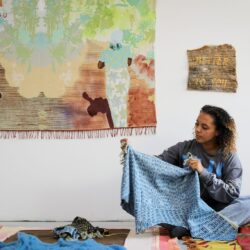 Malaika Temba sitting on a multicolored rug on the floor, holding a blue patterned fabric, behind her are three of her works hanging on a white wall.