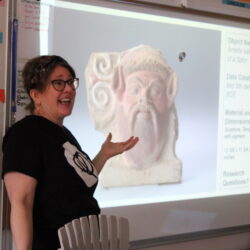 A woman in a classroom gestures toward an image projected on a smart board.