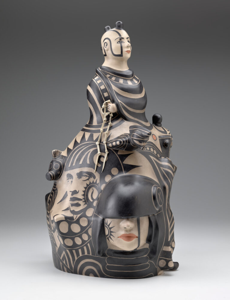Ceramic sculpture depicting a male bust at the top, with seven faces below, decorated in black and muted red-orange paint against a light beige base.