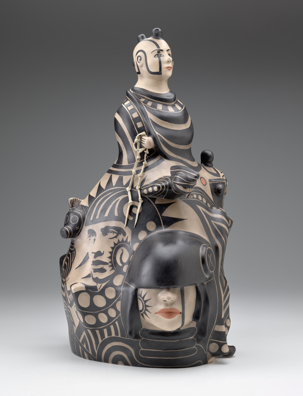 2023-09-18_ORTIZ_Convergence_Defenders_Descend_TR_2023_17_1_NA_view_2_PUB-1 Ceramic sculpture depicting a male bust at the top, with seven faces below, decorated in black and muted red-orange paint against a light beige base.