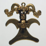 A symmetrical gold metal pendant of an eagle in flight with wings spread, wearing an ornate, two-headed snake headdress.