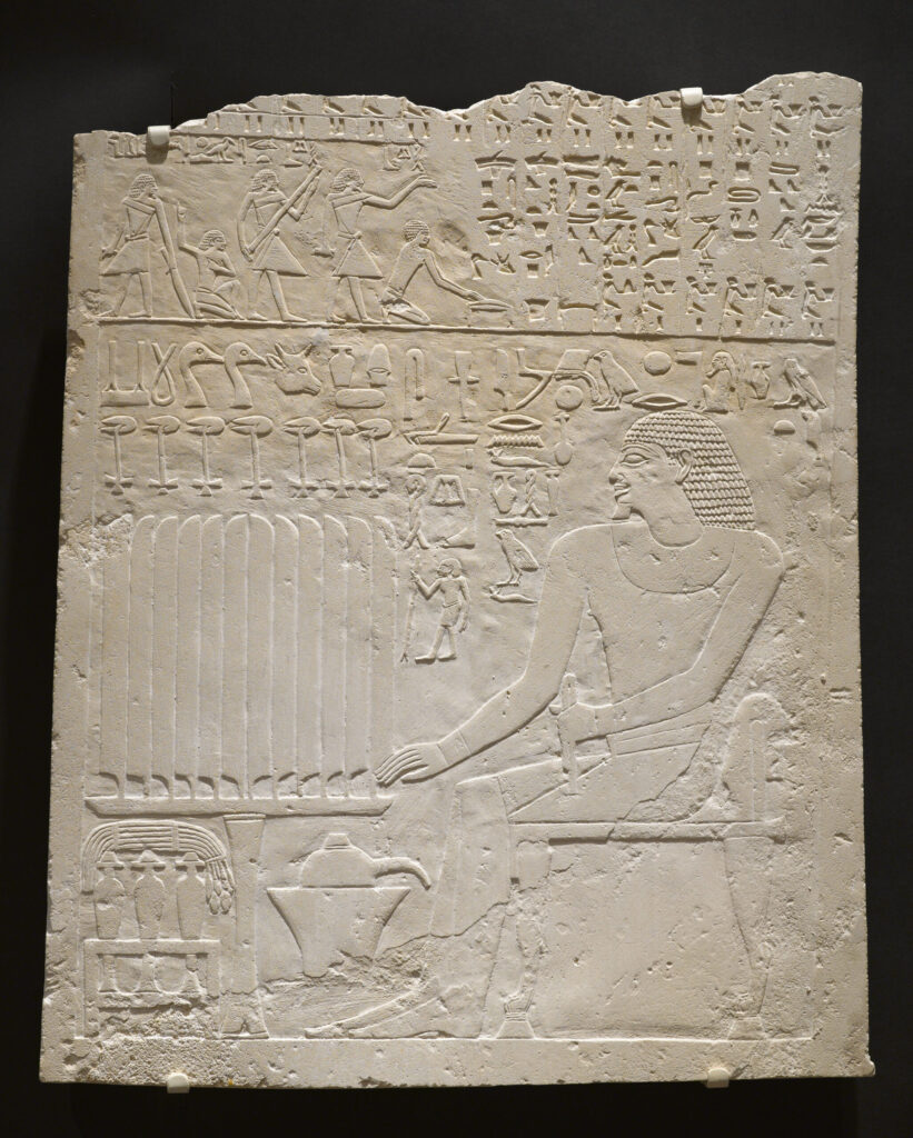 Carved limestone relief sculpture with jagged top edge, showing a seated man facing left, surrounded by hieroglyphs and small offering figures.