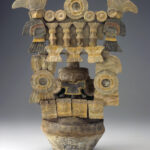 A ceramic incense burner with stacked architecture, circular motifs, and central mask-like face. A bird figure with wavy feathers perches atop each side.