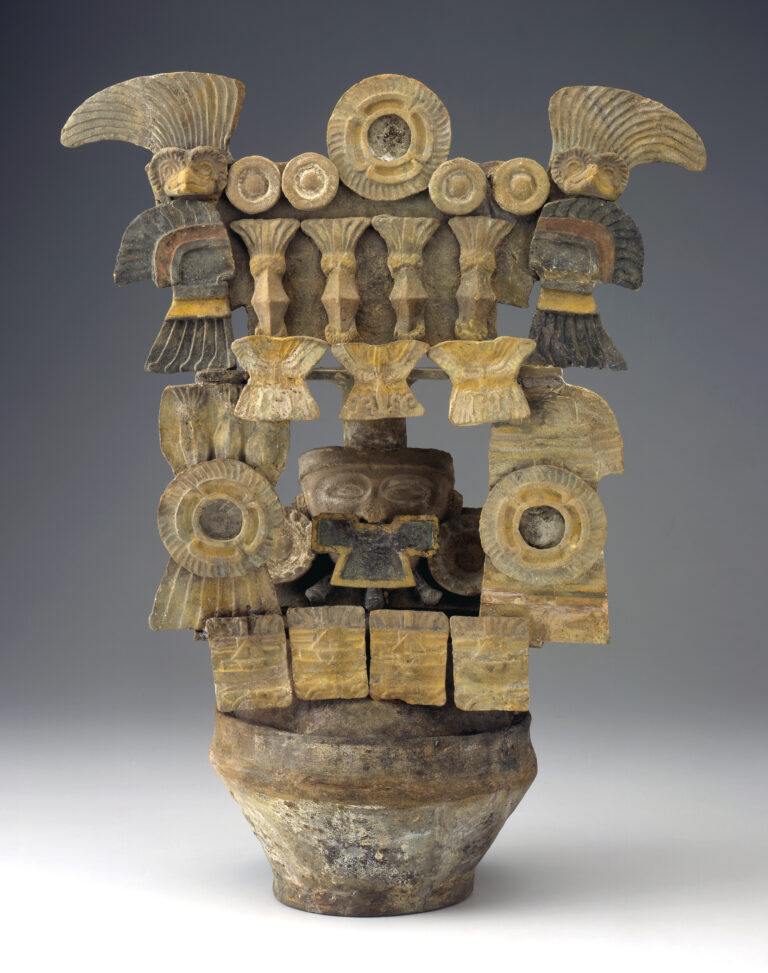 A ceramic incense burner with stacked architecture, circular motifs, and central mask-like face. A bird figure with wavy feathers perches atop each side.