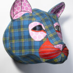 A mixed-media sculpture of a lion’s head made from blue plaid and red-striped cloth. Its facial features are accented with pink circle-patterned paper.