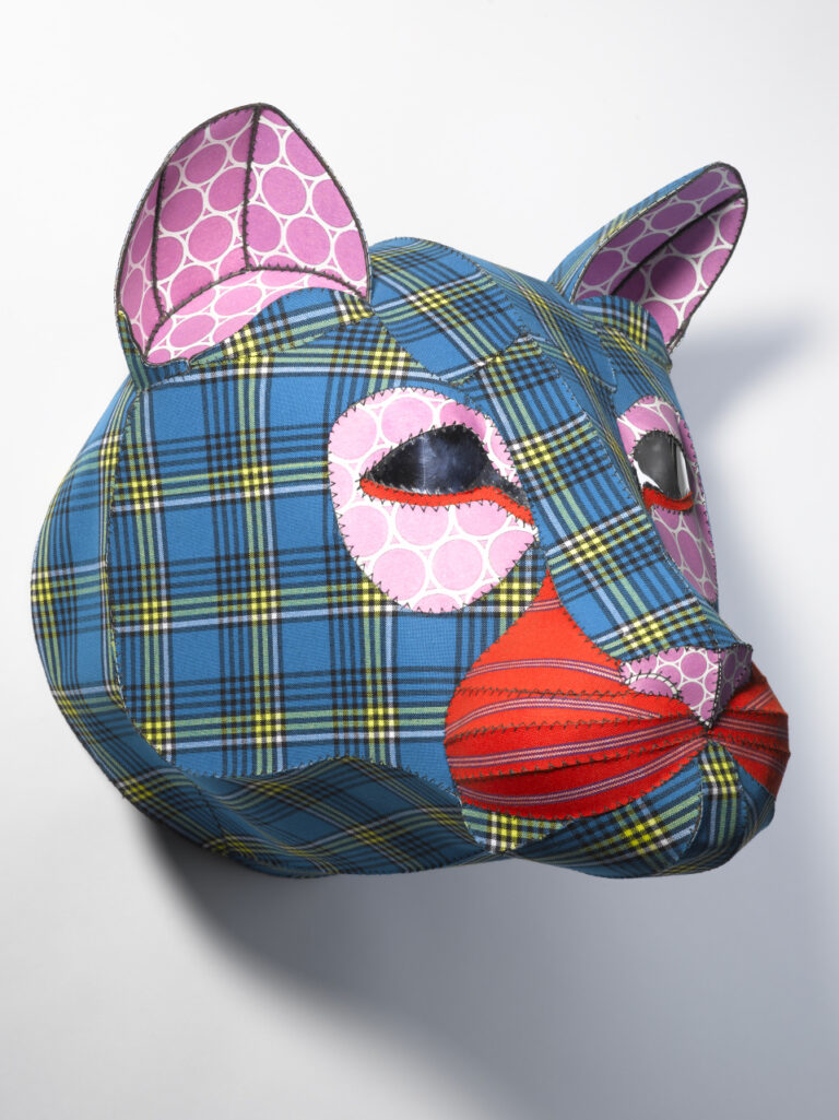 A mixed-media sculpture of a lion’s head made from blue plaid and red-striped cloth. Its facial features are accented with pink circle-patterned paper.