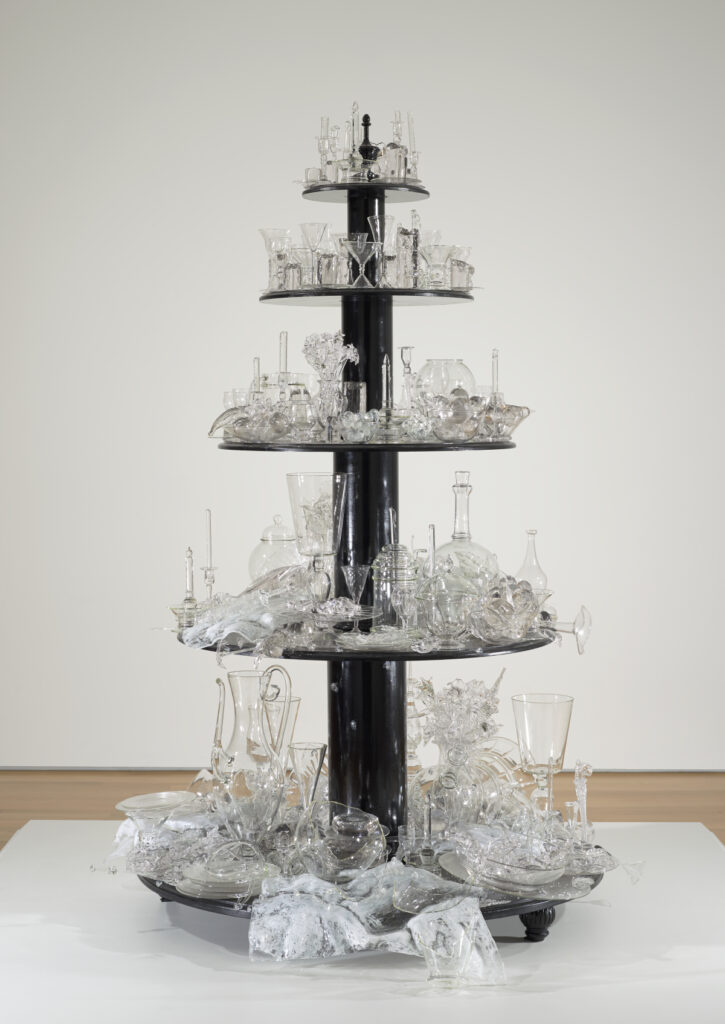 Sculpture made of five round wooden tiers painted black; each holds glass objects ranging from completely intact to broken and shattered.