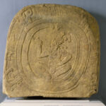 A tan, carved stone marker with a central design depicting a squatting man facing left.