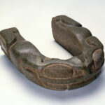 A carved u-shaped stone yoke resembling a frog with large eyes, a broad mouth at the front, and legs carved on the sides.