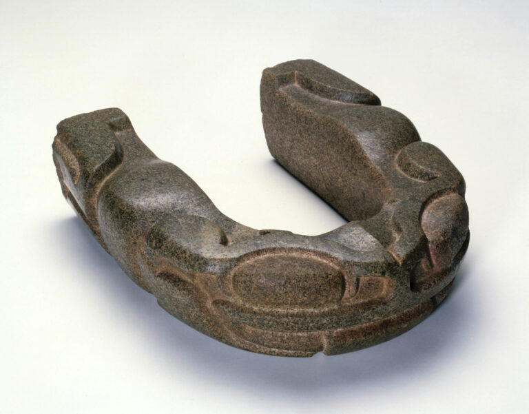 A carved u-shaped stone yoke resembling a frog with large eyes, a broad mouth at the front, and legs carved on the sides.