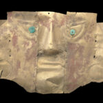 A hammered gold metal mask resembling a human face with blue-green eyes, prominent nose and lips, round earpieces, and traces of red pigment on the surface.