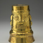 A gold cup positioned upside down, decorated with human facial features including eyes, a nose, open mouth, and ears.