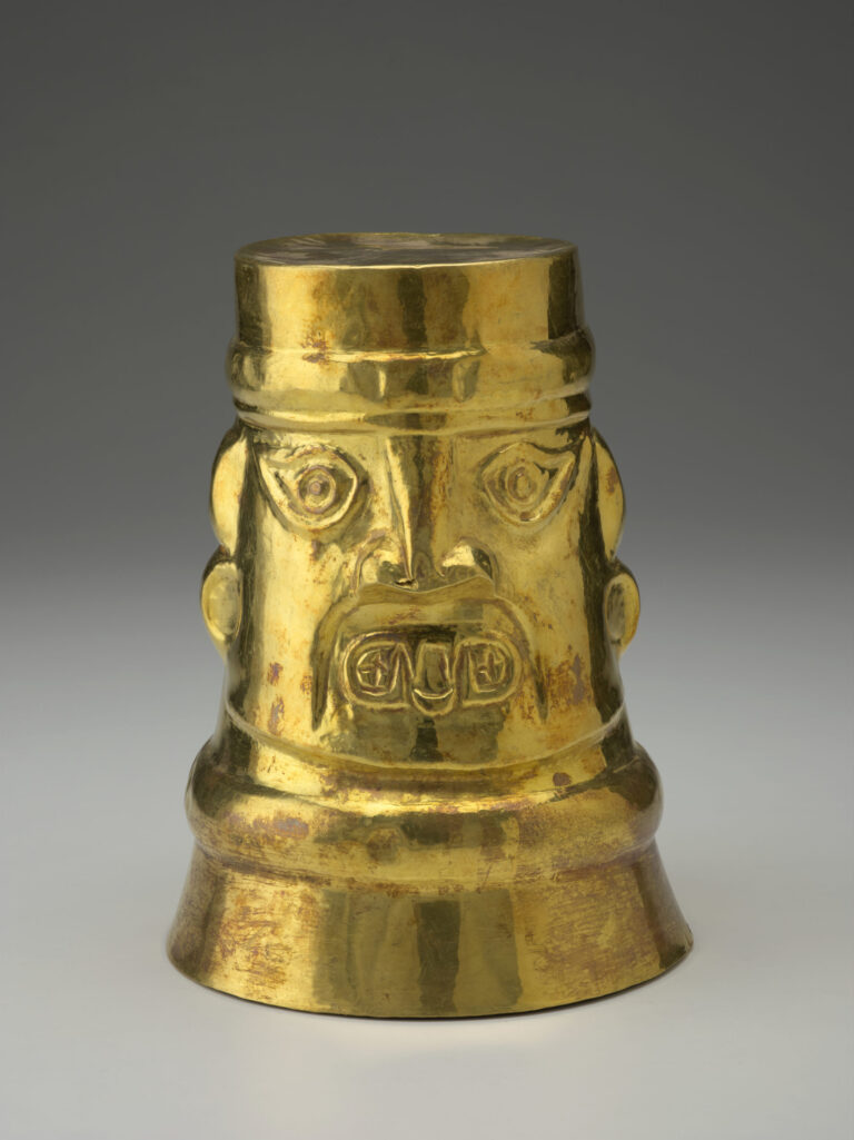 A gold cup positioned upside down, decorated with human facial features including eyes, a nose, open mouth, and ears.