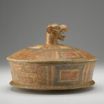 A terracotta cylindrical dish with a lid that has a jaguar-head handle. The dish is decorated with red and black designs.