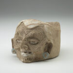 A light tan clay jar with a humanlike wrinkled face, eyes facing forward, and silver painted earrings on each ear.