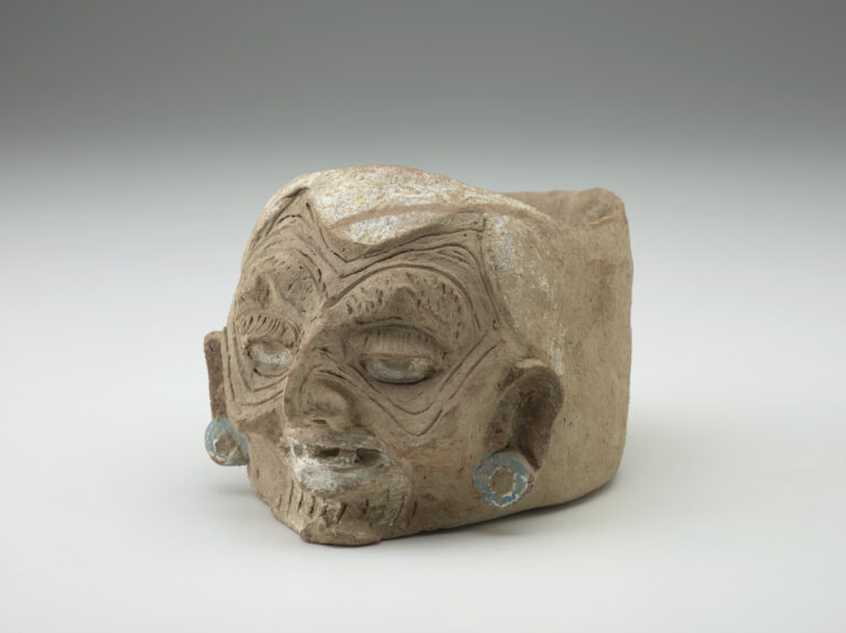 A light tan clay jar with a humanlike wrinkled face, eyes facing forward, and silver painted earrings on each ear.